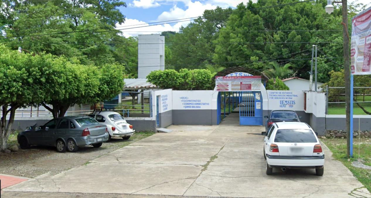 Fotografía en la que se muestra una explanada y al fondo se muestra la entrada al plantel, conformada por una reja de color azul y muros de color blanco con gris a los lados. Sobre la parte blanca de los muros, con letras azules están escritos algunos datos del plantel, como el nombre y las carreras que imparten. 